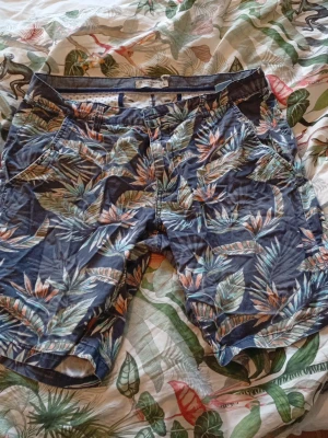 Shorts Iconic Port L - Säljer ett par blå shorts från Iconic Port i storlek L med tropiskt blommigt mönster i grönt, orange och beige. De har normal passform, dragkedja och knapp framtill samt fickor både fram och bak. Perfekta för varma dagar och chill häng. Men shortsen är en lite mindre än L tror jag så jag skulle snarare säga att det är i storleken M. Pris kan sänkas vid en villig affär 