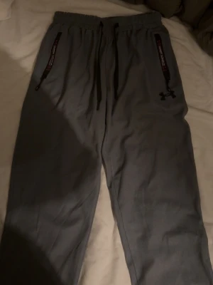 Grå mjukisbyxor från Under Armour - Grå mjukisbyxor från Under Armour med svarta snören i midjan och dragkedjor vid fickorna. Byxorna har ett löst passform och är tillverkade i ett syntetmaterial med ribbad struktur. Loggan syns tydligt på benet och detaljerna är svarta och röda.