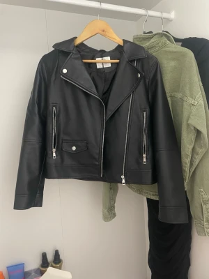 Svart skinnjacka med dragkedjor - Cool svart skinnjacka från Zara med klassisk bikerstil. Jackan har silvriga dragkedjor, bred krage och detaljerade fickor framtill. Perfekt för dig som vill ha en edgy look och gillar snygga detaljer. Passar dig som vill sticka ut lite extra.