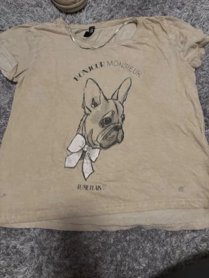 Beige t-shirt med fransk bulldog - Beige t-shirt från Colours of the World i storlek S. Framsidan har ett tryck med en fransk bulldog, stor vit rosett och texten 'Bonjour Monsieur' samt 'Tu me plais'. Rund hals och korta ärmar, mjukt bomullsmaterial. Perfekt för dig som gillar gulliga hundmotiv. Perfekt som pyjamas tröja 