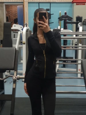 Svart slim träningsjacka med guldzip - Slimmad svart jacka med snygga guldiga dragkedjor framtill och på fickorna. Långärmad och figurnära passform, perfekt för dig som gillar sportig och stilren look. Materialet känns mjukt och stretchigt, vilket gör den bekväm att bära både till och från gymmet. Bara använd max 5 ggr. 