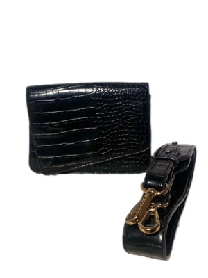 Svart axelväska med krokopräglat mönster - Snygg svart axelväska med krokopräglat mönster och guldfärgade detaljer. Väskan har ett justerbart axelband och magnetlås framtill. Insidan har flera fack, inklusive ett med dragkedja. Perfekt för dig som gillar stilrena accessoarer med lite edge.