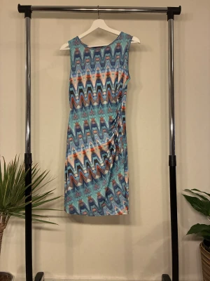 Mönstrad kortklänning - Färgstark kortklänning från Sisters Point. Klänningen har ett coolt zigzag-mönster i blått, orange och beige toner, är ärmlös och har en lätt rynkning på sidan. Perfekt för dig som gillar unika prints och vill sticka ut. OANVÄND 