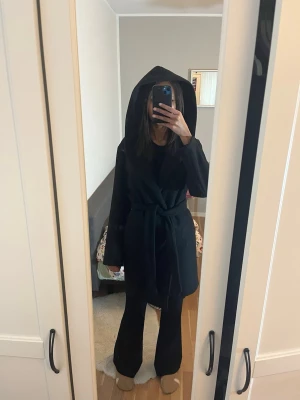 Svart kappa med huva och bälte - Säljer en svart kappa med stor huva och knytbälte i midjan. Jackan har lång ärm och en stilren, loose passform som ger en cool vibe. Perfekt för dig som gillar minimalistisk stil och vill ha något som sticker ut lite extra.