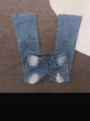 Blå bootcut jeans med slitningar - Snygga blå bootcut jeans med slitningar och ljusare tvättade partier fram och bak. Materialet är jeans och passformen är loose med utsvängda ben. Storlek 36. Midjemått 36cm rakt över innebenslängd 73cm
