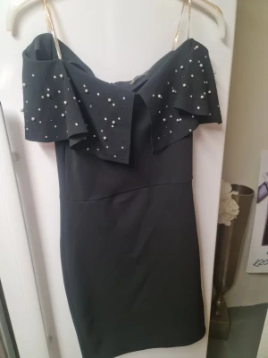 Svart kort klänning med pärldetaljer - Snygg svart kort klänning med axelbandslös design och dekorativa pärlor på volangdetaljen upptill. Klänningen har en figurnära passform och är perfekt för dig som vill sticka ut med stilrena detaljer. Materialet känns mjukt och har en lätt struktur.