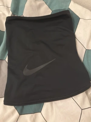 Svart Nike halsvärmare - Svart halsvärmare från Nike med diskret logga framtill. Tillverkad i mjukt och stretchigt syntetmaterial som känns skönt mot huden. Perfekt för kalla dagar och sportaktiviteter utomhus.