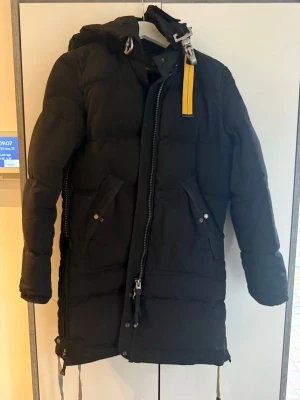 Svart Parajumpers lång pufferjacka  - Svart lång pufferjacka från Parajumpers med huva, gul detalj vid halsen och patch på axeln. Jackan har två stora fickor med knappar, dragkedja framtill och justerbara snören nedtill. Slim fit och riktigt varm för kalla dagar. Finns även päls till.
