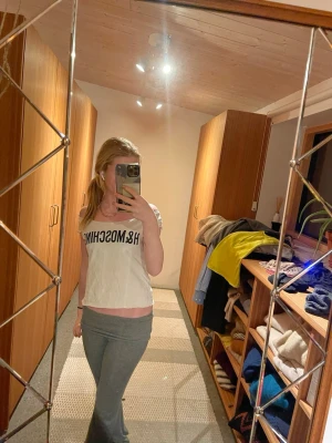 Vit croppad t-shirt med tryck och cool tyg design  - Säljer en vit croppad t-shirt från Moschino med svart tryck framtill. Toppen har offshoulder-design och coola utskurna detaljer i ryggen som ger en edgy vibe. Perfekt för dig som vill sticka ut och gillar streetstyle.