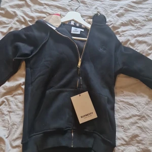 Svart hoodie från Burberry med dragkedja - Svart hoodie från Burberry med klassiskt rutigt foder i huvan och guldfärgad dragkedja. Tröjan har fickor framtill, långa ärmar och en broderad logga på bröstet. Perfekt för dig som vill ha en stilren och lyxig look.