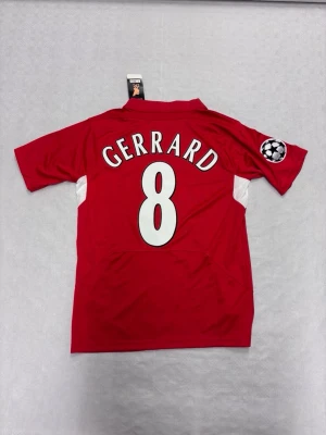 GERRARD Liverpool Hemma kit 04/05 - En röd Liverpool-tröja från Champions League-finalen 2005 med Gerrard och nummer 8 på ryggen, tillverkad av Reebok. Tröjan har Carlsberg-logga och broderade detaljer. Perfekt julklapp till ett liverpool fan.