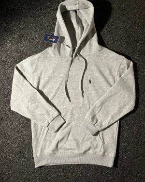 Grå hoodie från Polo Ralph Lauren - Snygg grå hoodie från Polo Ralph Lauren med klassisk logga broderad på bröstet. Tröjan har huva med snörning, magficka och ribbade muddar vid ärmslut och nederkant. Tillverkad i mjuk bomullsmix som är skön att bära. Perfekt för chill dagar.