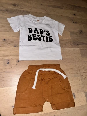 Bruna shorts med snörning för barn Set med T-shirt och Shorts - Säljer ett par bruna shorts för barn med vit snörning i midjan och praktiska fickor. Shortsen har en avslappnad passform och är perfekta för varma dagar. Tillverkade i mjukt bomullstyg för extra komfort.Ett sött set med en vit t-shirt med texten ”Dad's Bestie” och ett par bruna shorts med dragsko. Från cirka 6 mån storlek 