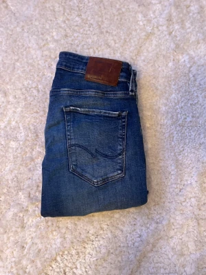 Blå Jack & Jones jeans slim fit - Snygga blå Jack & Jone jeans med slim fit och klassisk femficksdesign. Jeansen har en mörk tvätt med slitningar och detaljerade sömmar på bakfickorna samt läderpatch i midjan. Perfekt för dig som gillar en modern och avslappnad stil.