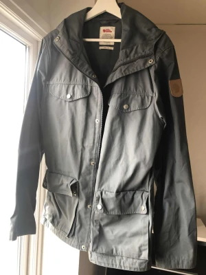Grå vindjacka från Fjällräven M - Snygg grå vindjacka från Fjällräven i storlek M. Jackan har huva, flera fickor med knappar och dragkedja framtill. Klassisk Fjällräven-logga på ärmen och stilren design som funkar till streetwear. Materialet är slitstark polyester och bomull.