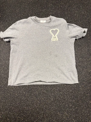Grå t-shirt från Ami Paris med hjärtdetalj - Snygg grå t-shirt från Ami Paris med klassisk hjärtdetalj och A-logga på bröstet. Klassisk passform, rund hals och korta ärmar. Tillverkad i mjuk bomull som känns skön mot huden. Säljer den då jag tycker att den är för liten på mig och får ingen användning av den.