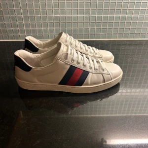 Gucci vita sneakers med blå och röd rand - Säljer ett par klassiska Gucci sneakers i vitt skinn med blå och röd rand på sidan. Baktill har de en mörkblå detalj med Gucci-logga och mönstrad struktur. Skorna har rund tå, snörning och platt sula med Gucci-mönster undertill. Perfekt för dig som gillar lyxiga och stilrena sneakers.