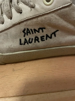 Saint Laurent vita sneakers med snörning - Det mesta kan nog gå och tvättas bort. Säljer ett par vita Saint Laurent sneakers med handskriven logga på sidan och guldfärgad text på plösen. Skorna har rund tå, platt sula och snörning. Ovandelen är i canvas och skinn, med slitna detaljer och råa kanter för en edgy vibe.