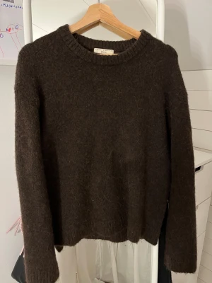 Brun stickad tröja från Gina - Mysig brun stickad tröja från Gina med rund hals och långa ärmar. Tröjan har en enkel och klassisk design med ribbade muddar vid hals, ärmslut och nederkant. Perfekt för kyliga dagar och enkel att matcha med jeans eller kjol.