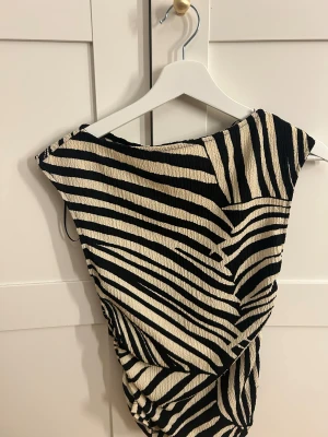 Zebramönstrad ärmlös topp - Säljer en cool ärmlös topp med zebramönster i svart och beige. Toppen har en lätt rynkad effekt på sidan och är tillverkad i ett strukturerat syntetmaterial. Perfekt för dig som gillar djurmönster och vill sticka ut med din stil.