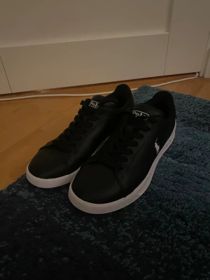 Svarta sneakers Polo Ralph Lauren Heritage Court II - Snygga svarta sneakers från Polo Ralph Lauren med vit logga på sidan och vit sula. Skorna har klassisk rund tå, svarta skosnören och är gjorda i skinnimitation. Perfekta för dig som gillar stilrena och tidlösa sneakers med en sportig vibe. Endast använda tre gånger och är i superfint skick. Nypris ca 1600kr. Perfekt julklapp!