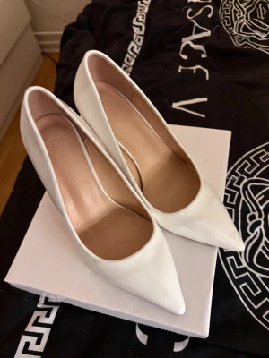 Vita pumps med röd sula och spetsig tå - Snygga vita pumps med glansig finish och röd undersida. Skorna har spetsig tå och hög klack (8-10 cm) Insidan är beige och materialet är syntet med skinnliknande känsla. Perfekt för dig som vill sticka ut med stil. Dom är endast använda inomhus 1-2 gånger. OBS se alla bilder!!