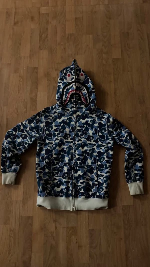 BAPE ZIP-UP hoodie  - Säljer en ikonisk BAPE shark hoodie i blått camo-mönster med dragkedja hela vägen och huva med hajansikte och broderade bokstäver. Tröjan har ribbade muddar och är tillverkad i mjuk bomull. Perfekt för dig som vill sticka ut med streetwear.