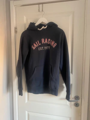 Marinblå Sail Racing hoodie - Säljer denna marinblå hoodien från Sail Racing. Hoodien är i mycket bra skick förutom ena snöret som slitits ut (sista bilden). Snörena går dock lätt att byta ut! Skriv vid minsta fråga 😁