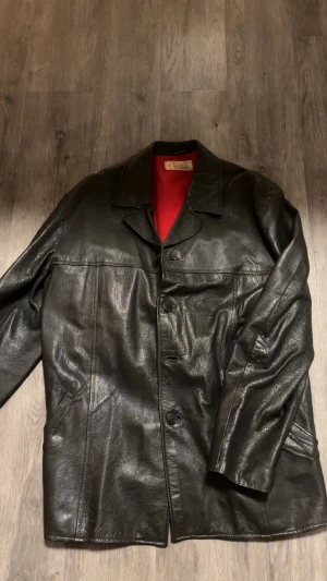 Svart skinnjacka från Äkta Getskinn - Säljer en svart skinnjacka från Äkta Getskinn, tillverkad i Sverige. Jackan har klassisk krage, knäppning med tre knappar och två sidofickor. Insidan är fodrad med rött tyg som ger en snygg kontrast. Perfekt för dig som gillar stilren och tidlös look.