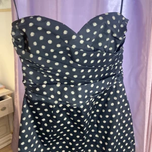 Svart prickig axelbandslös klänning - Snygg svart axelbandslös klänning med vita prickar och draperad överdel. Klänningen är kort och har en figurnära passform upptill med lätt utställd kjol. Perfekt för dig som vill sticka ut med ett lekfullt mönster och en klassisk siluett.tror den är oanvänd eller använd 1 gång passar för fest eller vardag mycket elegant.