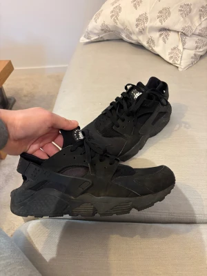 Nike Air Huarache svarta sneakers - Svarta Nike Air Huarache sneakers med chunky sula och klassisk snörning. Ovandelen är i en mix av syntet och mocka med detaljer i gummi runt hälen. Ikonisk Huarache-logga på plösen och rund tå. Perfekt för dig som gillar streetwear och sportig stil. Använda ett par gånger bara. 