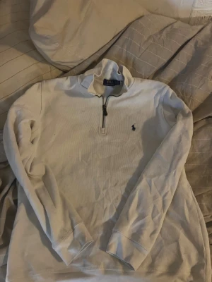 Vit half zip tröja från Polo Ralph Lauren - Vit långärmad tröja från Polo Ralph Lauren med klassisk half zip-dragkedja och broderad logga på bröstet. Tröjan är i bomull och har en stilren, enkel design som passar perfekt till jeans eller chinos.