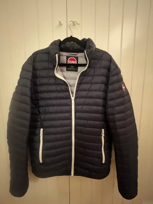 Marinblå pufferjacka från Colmar - Snygg Marinblå pufferjacka från Colmar med vit dragkedja och två sidofickor med dragkedja. Jackan har ett quiltat mönster och Colmar-logga på ärmen. Perfekt för kyliga dagar och har en modern, clean look. Perfekt all around jacka som man kan använda på hela året runt 🤩