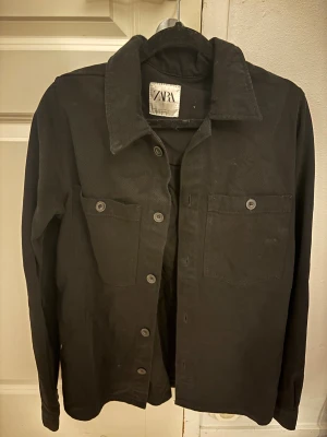 Svart overshirt från Zara - Stilren svart overshirt från Zara med klassisk krage, två bröstfickor med knappar och raka ärmar. Skjortan är tillverkad i bomull och har en enkel, clean look som passar till många outfits.