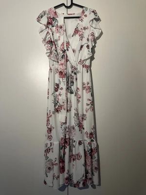Vit blommig klänning, storlek M - Jättefin vit klänning med blommigt mönster i rosa och grått. Klänningen har volanger vid axlarna och ett band som knyts i midjan.