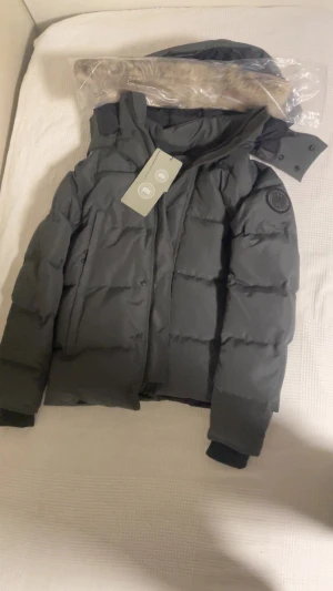 Svart dunjacka från Canada Goose - Snygg svart dunjacka från Canada Goose med huva och klassisk patch på ärmen. Jackan har dragkedja och knappar framtill, flera fickor och ribbade muddar vid ärmslut. Perfekt för kalla dagar och har en clean, stilren look.