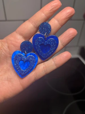 Blå glittriga hjärtörhängen - Snygga örhängen i akryl med intensivt blå färg och glitterdetaljer. Örhängena har en rund topp och ett stort hjärta med glittrande inramning. Perfekta för att sticka ut och ge din look en färgklick.