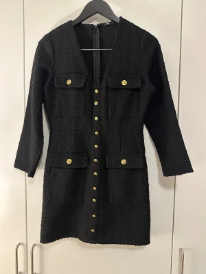 Svart kort klänning från Zara - Stilren svart kort klänning från Zara med markerad struktur, djup v-ringning och dekorativa guldknappar framtill. Fyra fickor med lock och trekvartsärm ger en chic look. Perfekt för dig som gillar klassiska detaljer och modern passform.