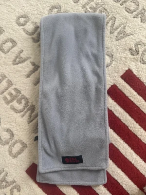 Ljusgrå fleecehalsduk Fjällräven - Mjuk och varm ljusgrå halsduk från Fjällräven i fleece. Rektangulär modell med måtten 130x20 cm. Perfekt för kalla dagar och enkel att matcha med olika outfits. Diskret svart etikett med röd logga på ena kanten.