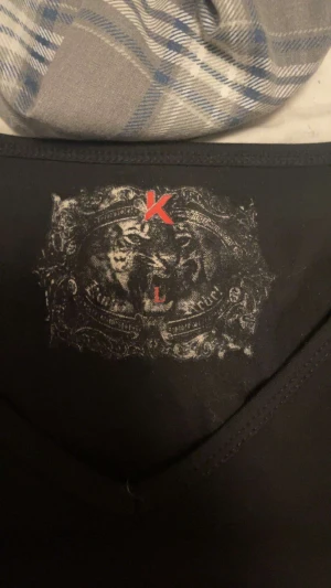 Svart t-shirt med kors och sköld - Svart t-shirt från KLG med stort tryck av röd sköld och kors i guld och svart på både fram- och baksida. T-shirten har korta ärmar och detaljer i guld, samt text och emblem som ger en edgy vibe. Materialet känns som bomull och passformen är normal.