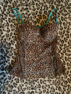 Leopardmönstrad korsettopp - Säljer en leopardmönstrad korsettopp från BASIC med vadderade kupor och justerbara turkosa axelband. Toppen har en liten turkos rosett framtill och dekorativa turkosa sömmar längs sidorna. Materialet är mjuk polyester med stretch för en tight passform. Kupa 36B.
