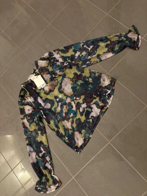 Blommig blus från ICHI med volangärm - Säljer en transparent blus från ICHI med färgstarkt blommönster i grönt, lila, blått och svart. Blusen har långa ärmar med volangdetalj vid ärmslut och en lätt glansig finish. Perfekt för dig som gillar statement-plagg och unika prints.