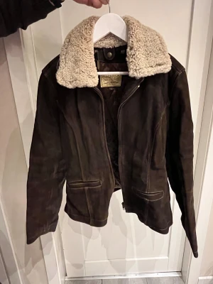 Brun skinnjacka med teddykrage Redskins - Snygg brun skinnjacka från Redskins med  avtagbar teddykrage i beige. Jackan har dragkedja framtill, två fickor och quiltat innerfoder. Klassisk modell med coola sömmar och detaljer, perfekt för dig som vill ha en unik look.