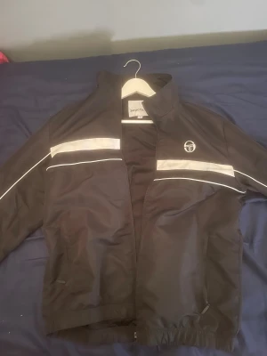 Svart vindjacka från Sergio Tacchini storlek M/L - Svart vindjacka från Sergio Tacchini med vita reflexdetaljer och logga på bröstet samt stor logotyp och text på ryggen. Jackan har dragkedja framtill, ståkrage och två sidofickor med dragkedja. Perfekt för dig som gillar sportig stil.