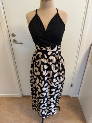 Svart och beige leopardklänning Shein - Snygg långklänning från Shein med svart omlottliv och smala axelband. Kjolen har ett beige och svart leopardmönster som ger en cool kontrast. Klänningen är tillverkad i polyester och har en figurnära midja med lätt rynk. Perfekt för dig som vill sticka ut.