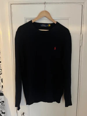 Mörkblå stickad tröja Polo Ralph Lauren - Snygg mörkblå stickad tröja från Polo Ralph Lauren i storlek M. Klassisk design med rund hals och röd broderad logga på bröstet. Tröjan har långa ärmar och ribbade muddar vid hals, ärmslut och nederkant. Perfekt för dig som gillar stilrena plagg. Vill få den såld därav de låga priset. Hör av dig vid frågor eller funderingar!!