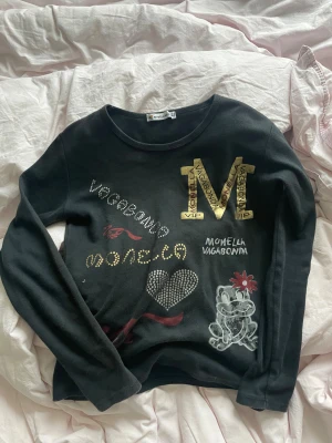 Svart långärmad tröja Monella Vagabonda - Svart långärmad tröja från Monella Vagabonda med coola tryck och detaljer i guld, silver och rött. Stor S i guld, strasshjärta, text och en söt groda med röd blomma framtill. Rund hals och mjukt bomullsmaterial. Perfekt för dig som gillar statement-plagg.