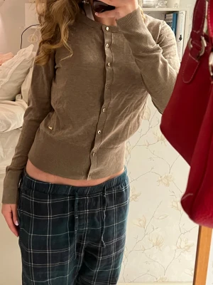 Beige kofta från Holly & White - Snygg beige kofta från Holly & Whyte by Lindex i storlek S. Den har rund hals, långa ärmar och guldfärgade knappar framtill. Materialet är mjukt och stickat, perfekt för lager på lager.  