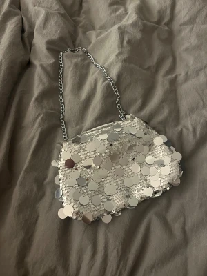 Silvrig paljettväska med kedja - Glittrig kuvertväska i silver. Väskan har en metallkedja som axelrem och stängs med dragkedja upptill. Perfekt för att addera lite extra bling till din outfit.