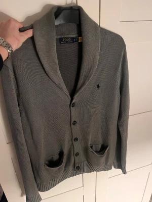 Grå kofta från Polo Ralph Lauren - Snygg grå kofta från Polo Ralph Lauren med klassisk sjalkrage och knappar framtill. Tröjan har två fickor och den ikoniska broderade loggan på bröstet. Stickad i mjukt material och har ribbade muddar vid ärmslut och nederkant. Perfekt för lager-på-lager.